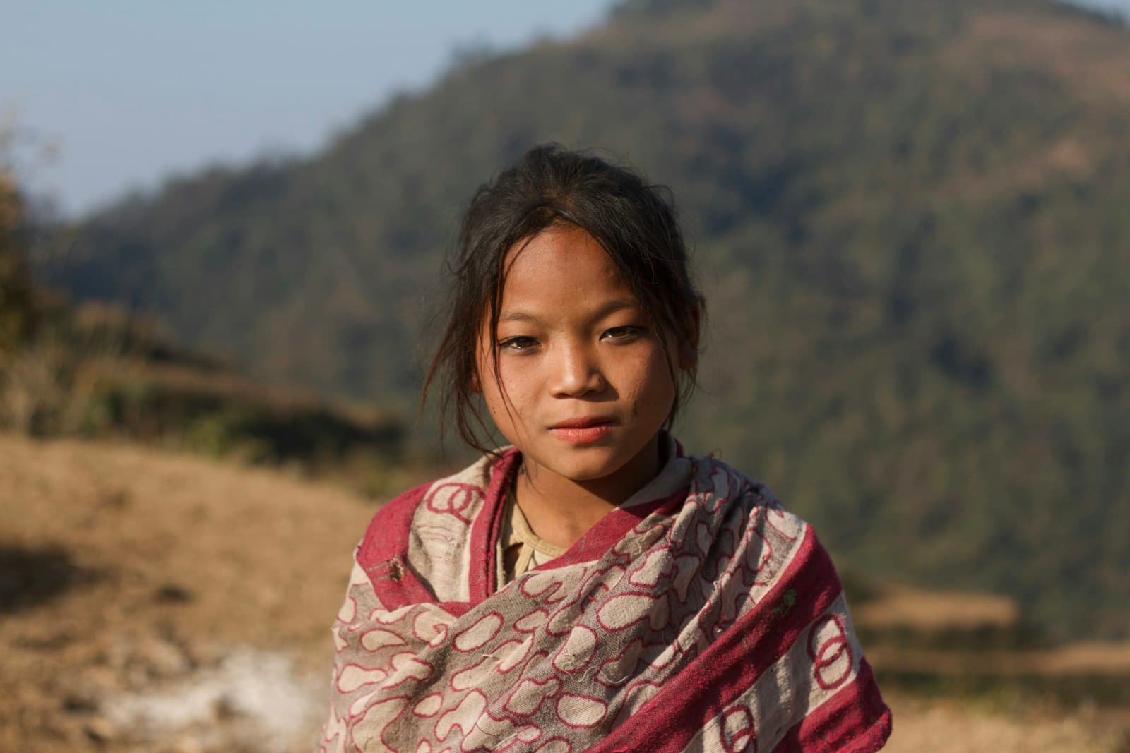 A young girl in a warm striped shawl before a softly blurred mountain hillside.